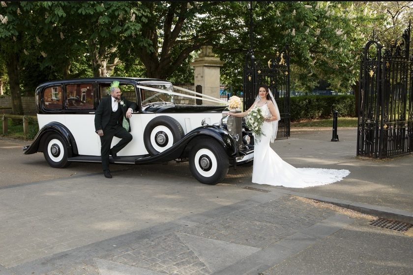 A bride and groom beside the black over white 1937 Phantom III by a pair of iron gates.