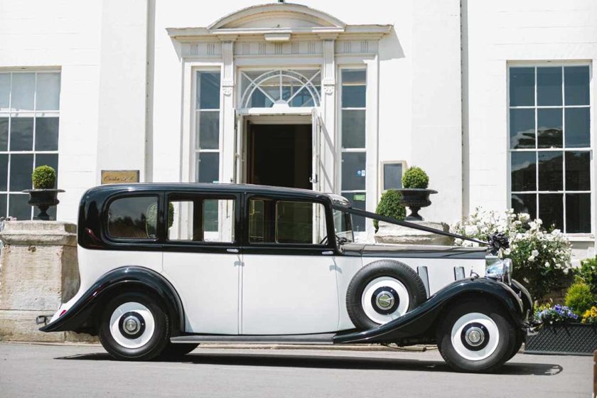 A black over white 1937 Phantom III vintage Rolls Royce outside a white Georgian hall.