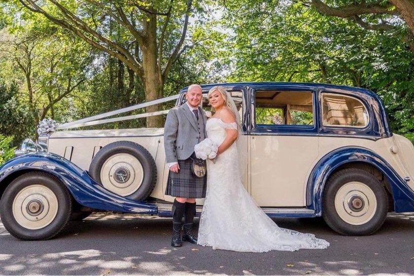A bride and groom, the groom in a kilt, beside the navy and cream 1938 Phantom III.