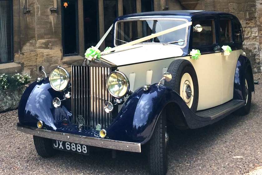 A vintage Rolls Royce in navy and cream, dressed with ivory ribbons, outside a stone building.