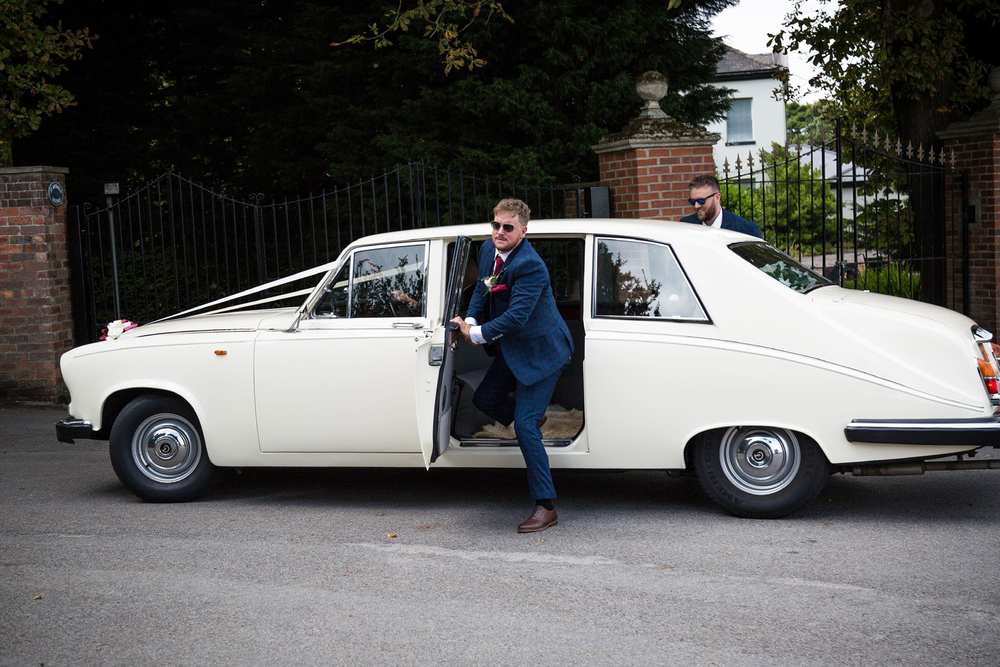 A groom stepping out of an ivory Daimler limousine while the chauffeur waits at the rear of the car.