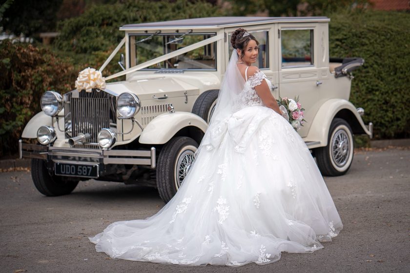 A bride in a lace ball gown looking over her shoulder in front of an ivory Imperial Viscount Landaulette dressed with ivory ribbons and a cream bow.