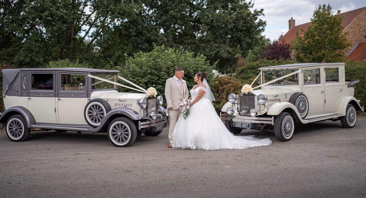 Two vintage-style Imperial Viscount wedding cars, one ivory and one ivory and silver, dressed with ribbons and cream bows, with a bride and groom standing between them.